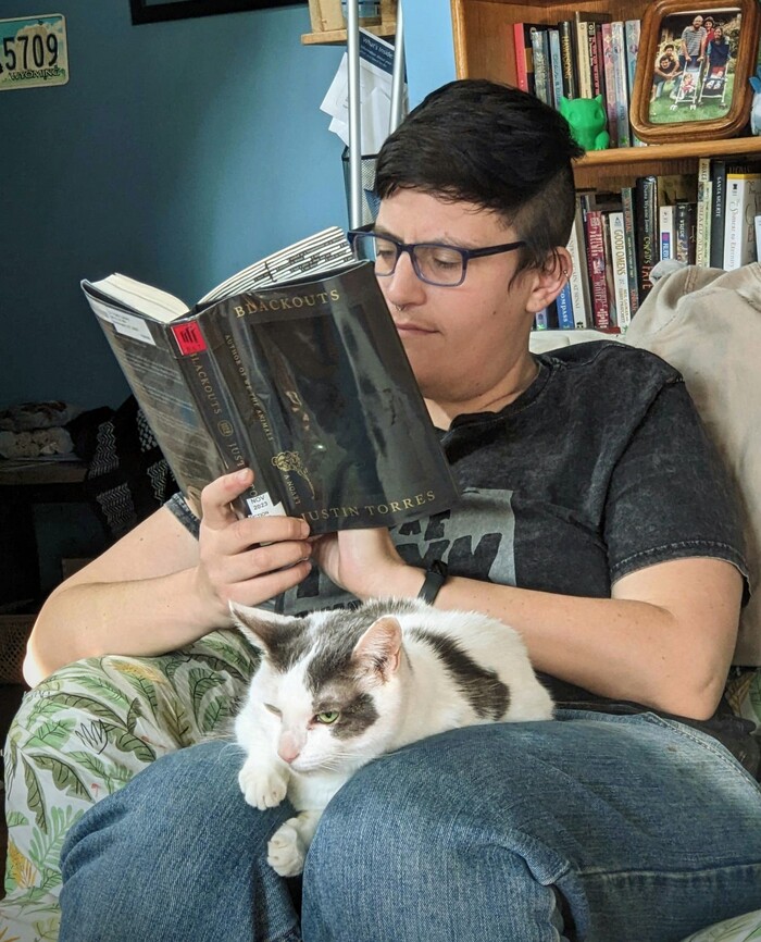 [photo of a white person with short dark hair sitting in a chair with a white and tabby cat on their lap, reading a library copy of 'Blackouts' by Justin Torres]